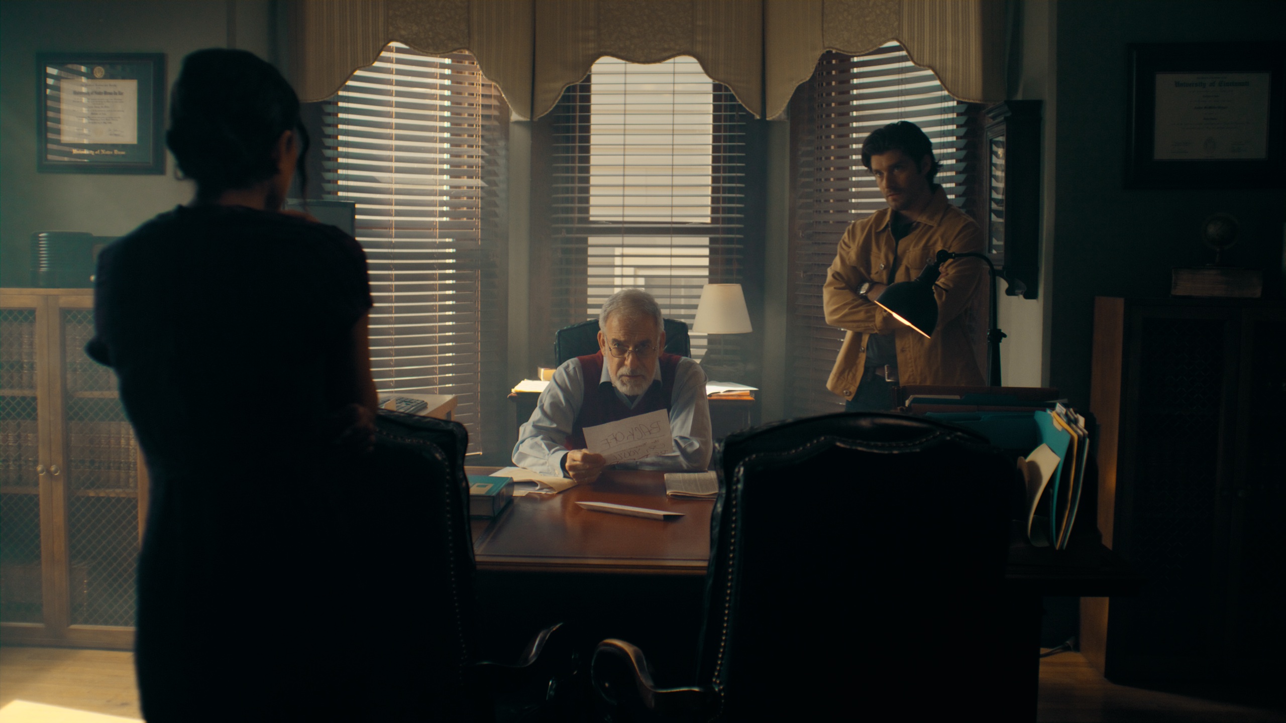Three figures in dark office with desk lamp and blinds