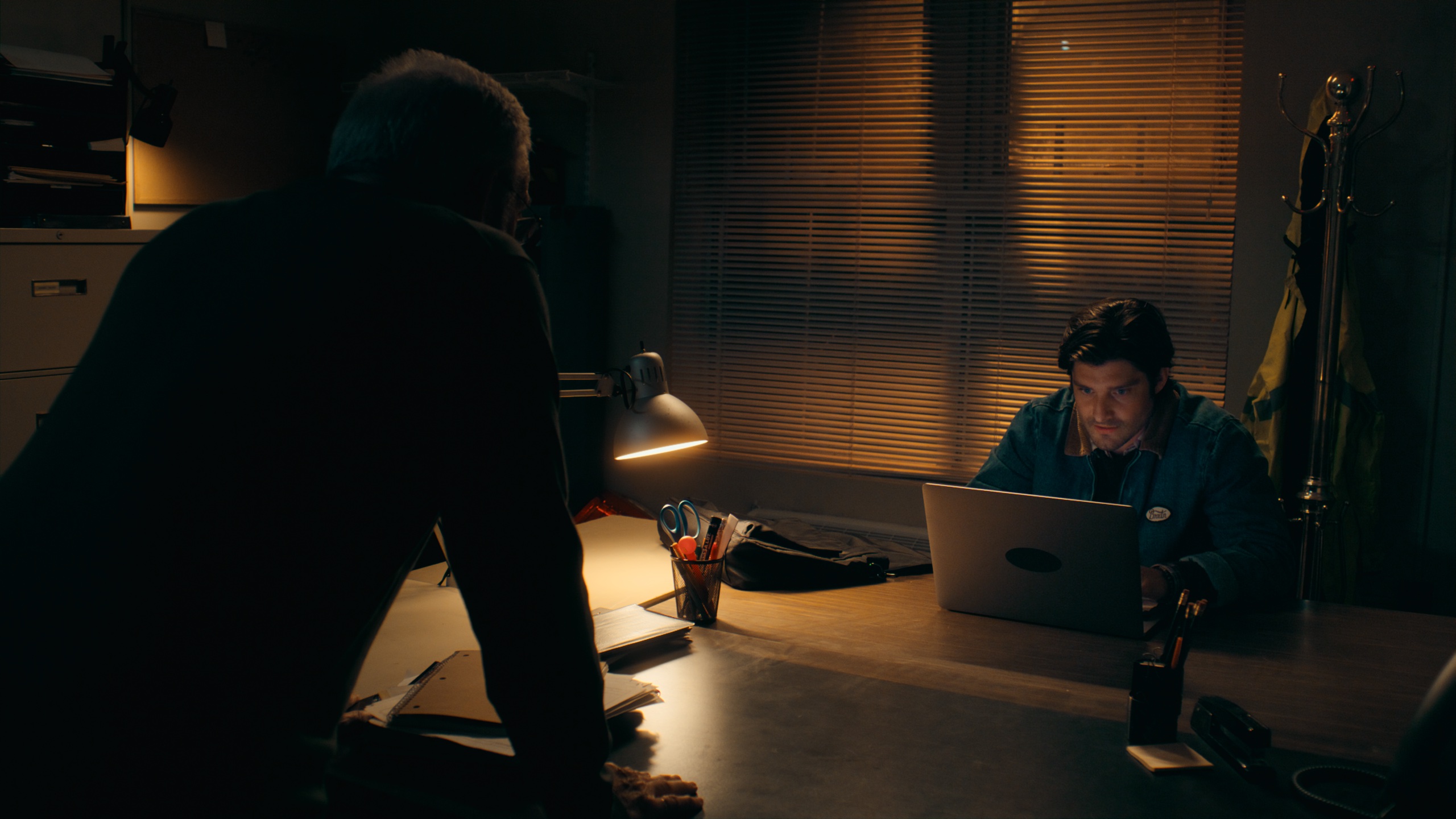 Two men in dark office lit by desk lamp and laptop